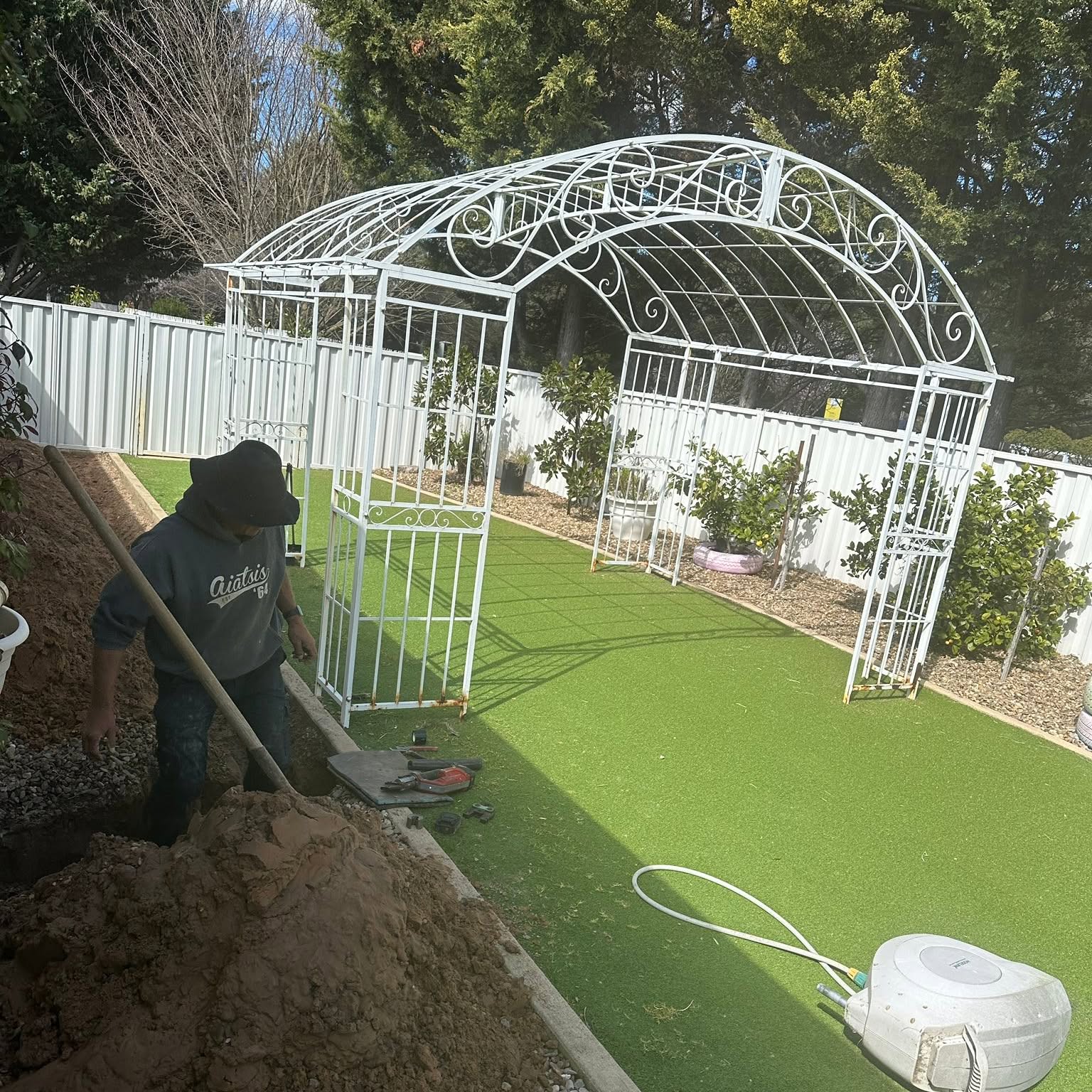 Digging out a service run beside a garden arbour, tools laid out on the lawn