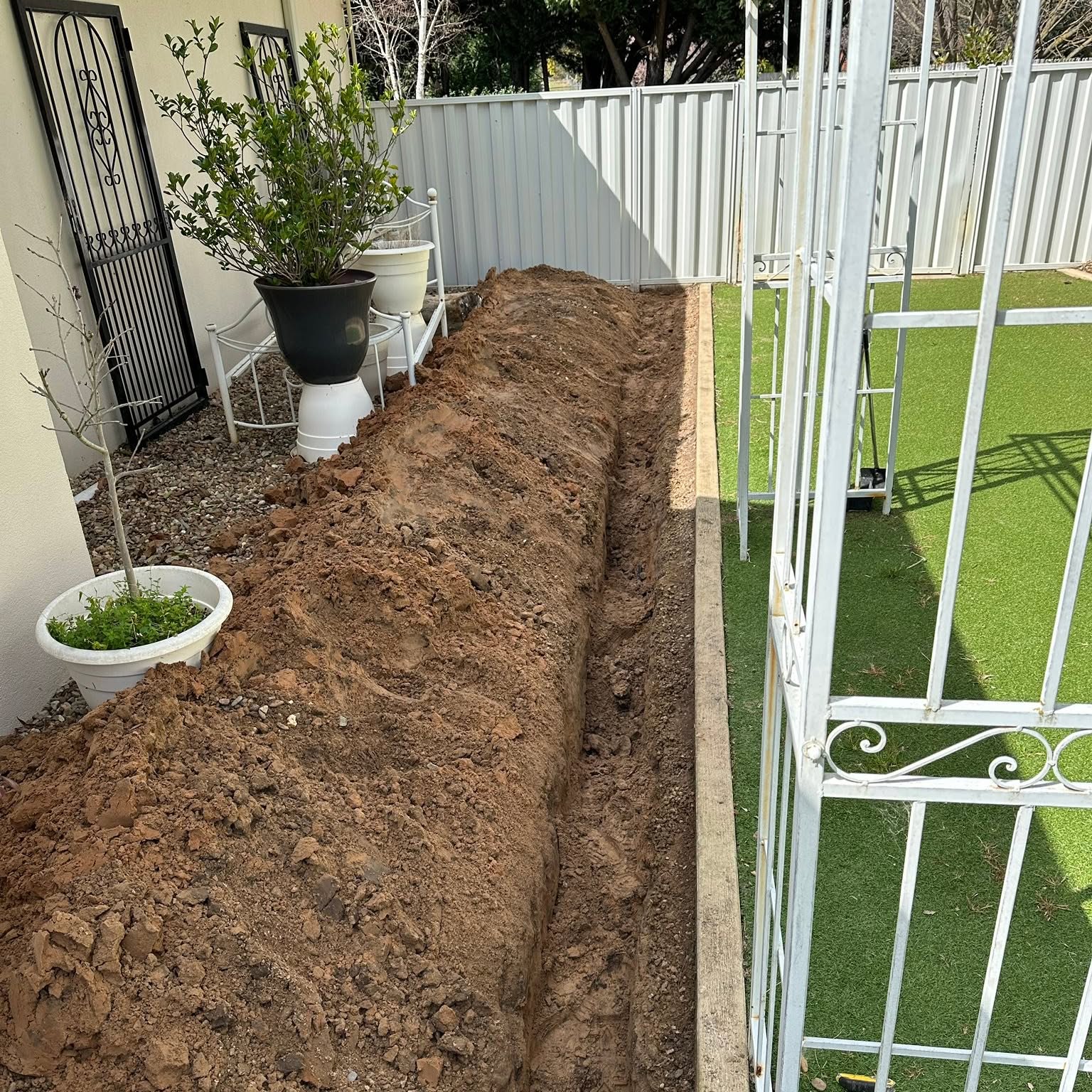 Long straight trench dug down the side of a house beside a garden bed